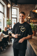 Mann steht im Café und trägt schwarzes Love Like Jesus Oversized T-Shirt mit goldenem Herz